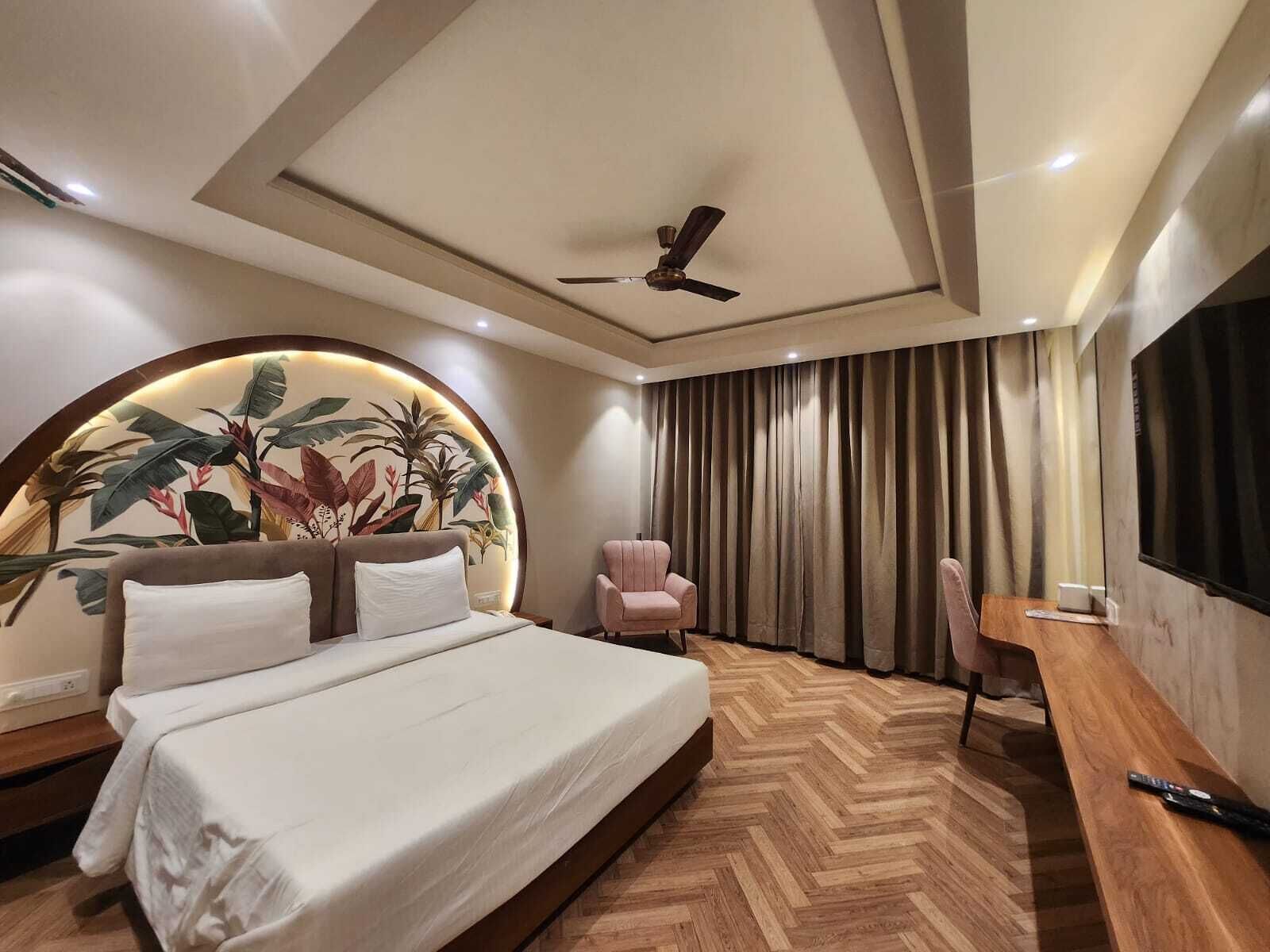 A modern hotel room with a large bed, a desk, a chair, a ceiling fan, and a wall-mounted TV.