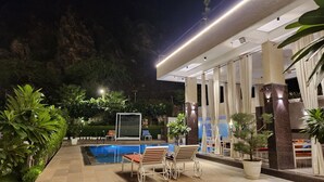 Uma área de piscina com espreguiçadeiras e trampolim, cercada por plantas em vasos e um muro de pedra.
