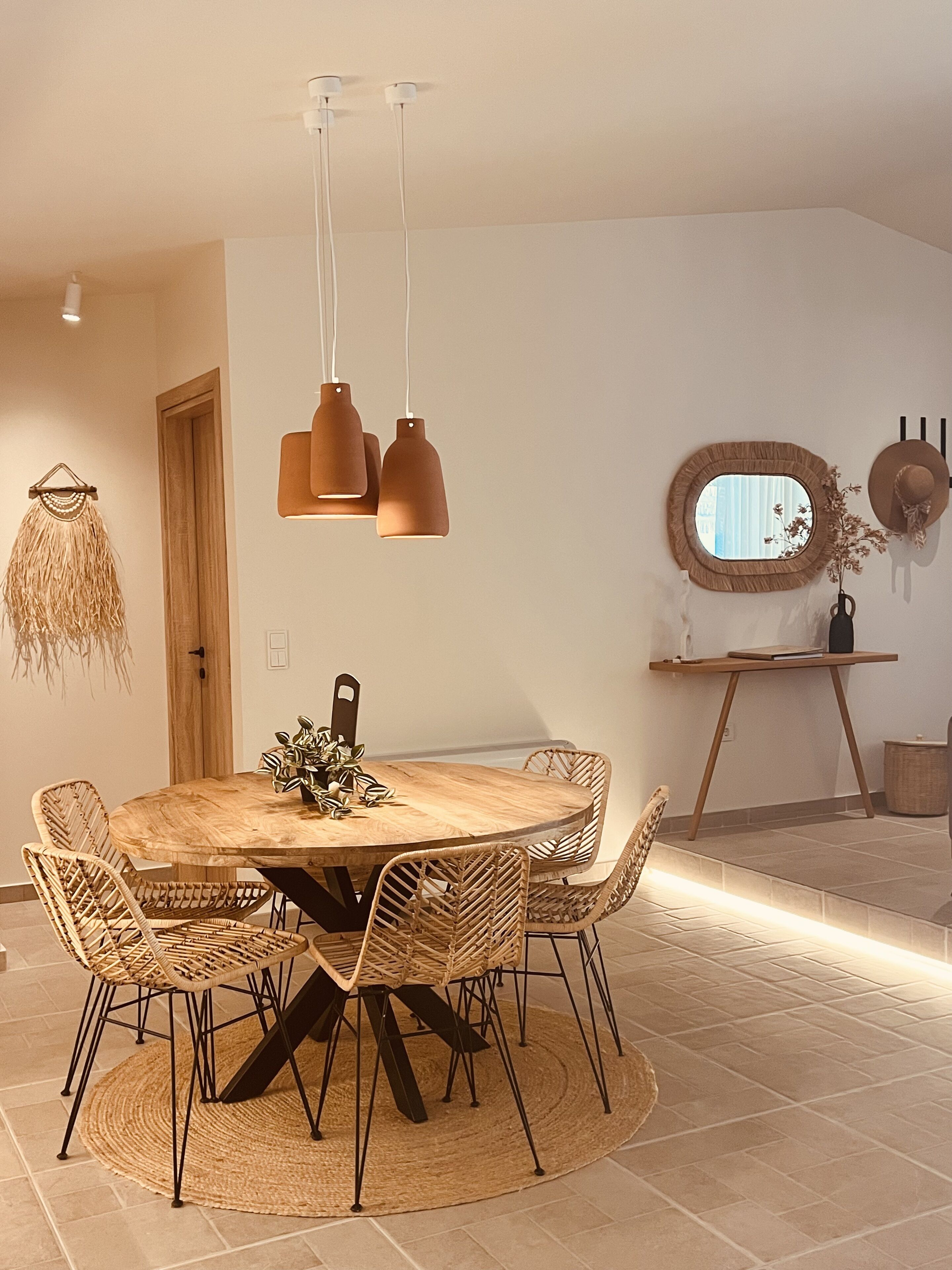Uma área de jantar com uma mesa redonda de madeira, rodeada por cadeiras de vime, um tapete tecido e luminárias pendentes.