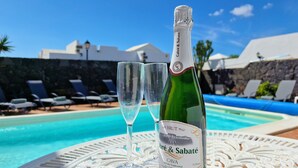 Una botella de cava Brut y dos copas sobre una mesa junto a una piscina, con sillones de descanso y un muro de piedra al fondo.