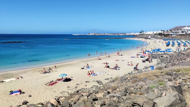 Una playa con gente tomando el sol y nadando, costa rocosa y edificios al fondo.