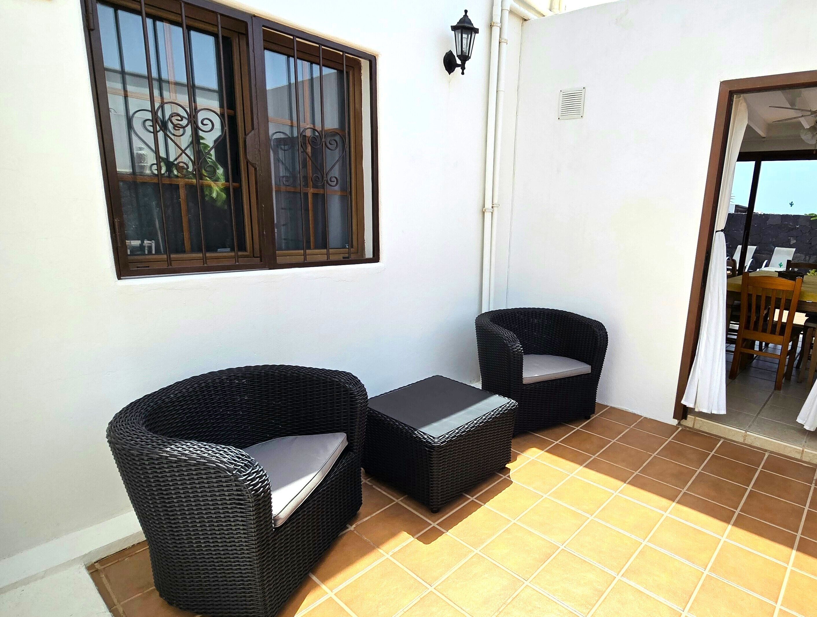 Un petit espace patio avec deux chaises en osier et une table, une fenêtre avec des barreaux et un luminaire fixé au mur.