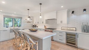 A modern kitchen with white cabinetry, a large island with bar stools, a built-in oven, and pendant lighting. - 3907 Twin Tides - Updated Beach House with Hot Tub (Carmel)