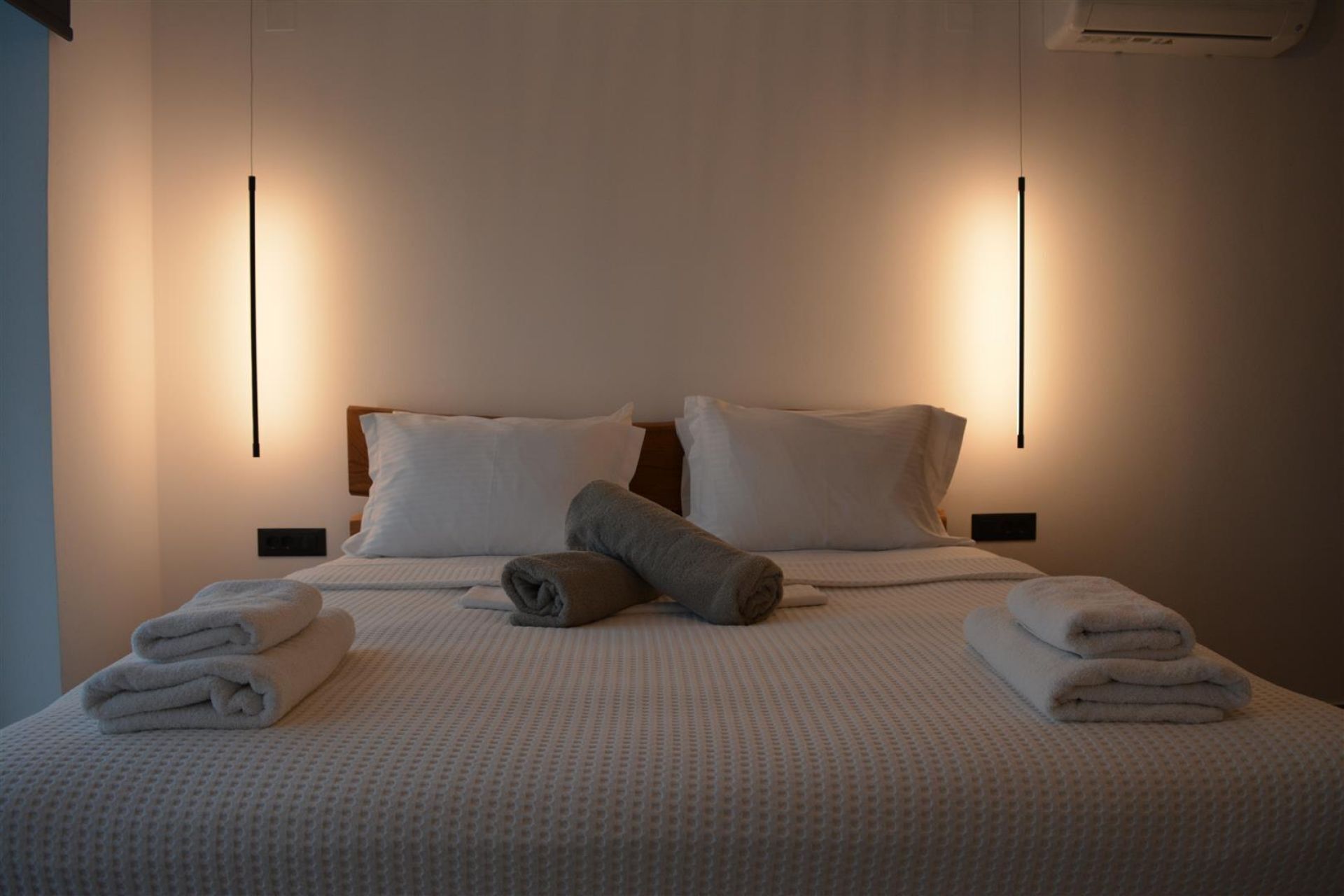 Una cama bien hecha con sábanas blancas y dos toallas enrolladas, un cabecero de madera y dos lámparas fijadas a la pared.