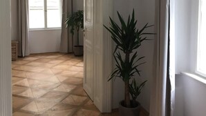 A spacious room with a wooden parquet floor, a large window with curtains, and a potted plant.