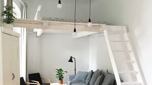 A compact living space with a loft bed, a sofa, a chair, a small table, and a potted plant.