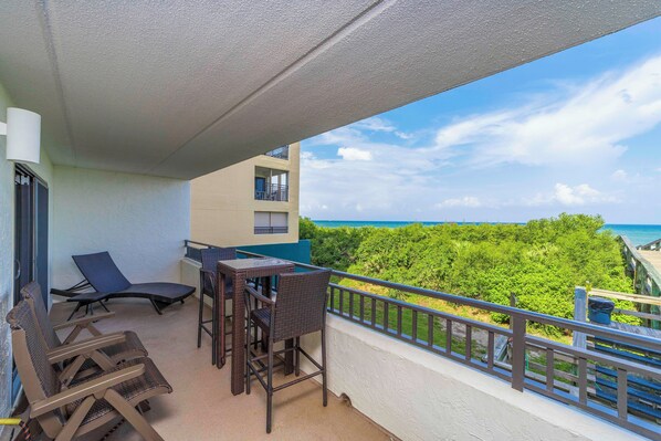 A balcony with wicker furniture, a table, and chairs, overlooking a lush green landscape and the sea.