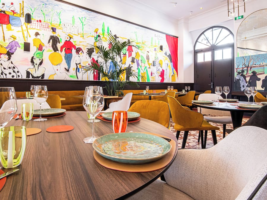 Un restaurante con mesas listas para comer, un mural grande y colorido en la pared y una planta en el centro.