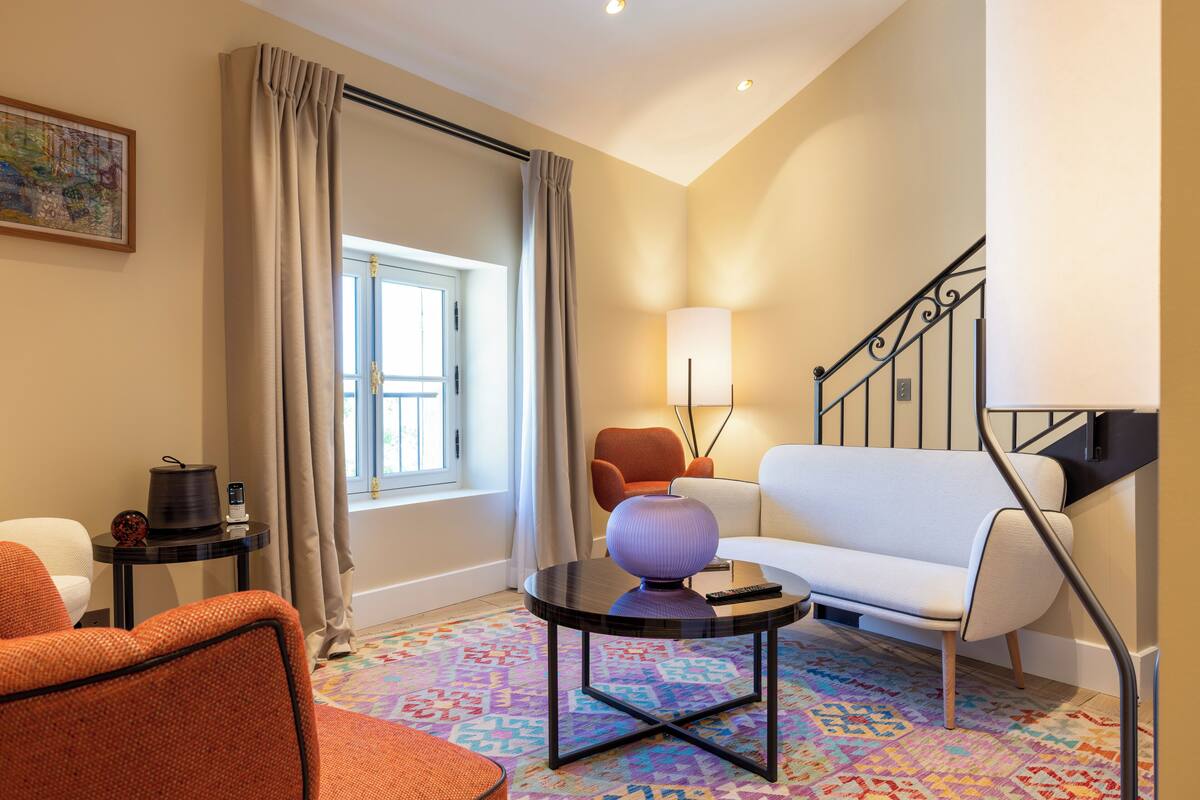 Habitació d'hotel moderna amb un sofà blanc, una butaca taronja i una taula de centre rodona amb una làmpada. Una catifa colorida recobreix el terra, i es veu una finestra amb cortines.
