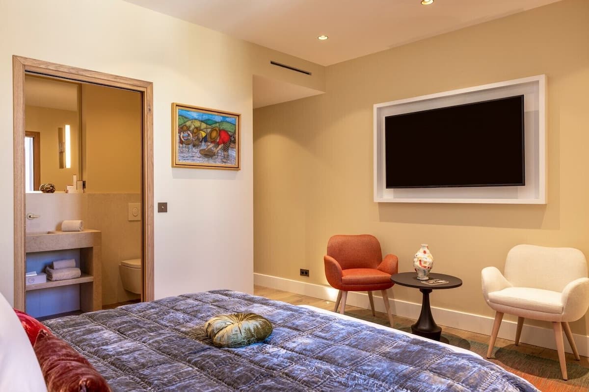 Habitació d'hotel amb un llit, dues cadires, una televisió, un quadre i un bany visible a través d'una porta oberta.