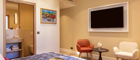 A hotel room with a bed, two chairs, a TV, a painting, and a bathroom visible through an open door.