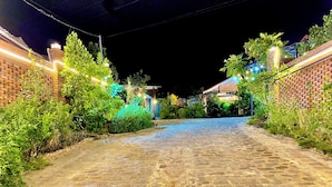Un chemin pavé, bordé de murs en briques et d’une végétation illuminée, mène à un bâtiment dont l’entrée est bien visible.