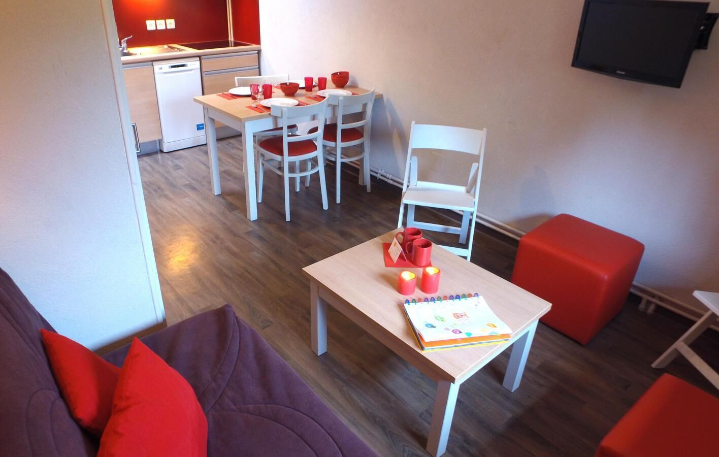 A modern living room with a sofa, a dining table with chairs, a small kitchen area, and a red ottoman.