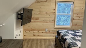 A bedroom with a sloped ceiling, wooden walls, a window, a bed, and a TV mounted on the wall.