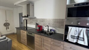 A modern kitchen with a built-in oven, microwave, and a coffee machine. There is a sink with a faucet, and a countertop with a kettle and a red coffee maker. The kitchen has wooden cabinets and a backsplash with patterned tiles.