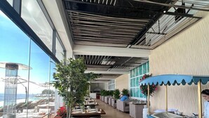 Ein Restaurant mit hohen Decken, großen Fenstern und Blick auf den Ozean. Die Tische sind mit Tellern, Gläsern und Besteck gedeckt.
