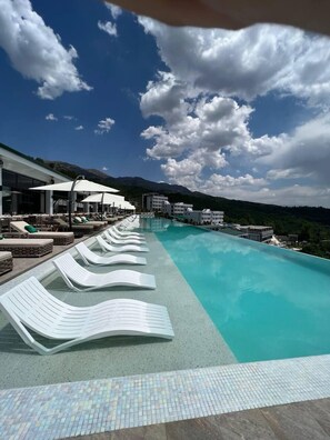 A poolside area with lounge chairs and umbrellas, a tiled walkway, and a view of mountains under a partly cloudy sky. - Heaven's Garden (Saylyk)