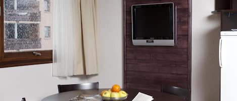 A compact living space with a round dining table, dark wooden chairs, a small TV mounted on a wooden panel, a white refrigerator, and a window with a view of buildings outside.