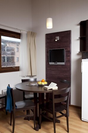 A compact living space with a round dining table, dark wooden chairs, a small TV mounted on a wooden panel, a white refrigerator, and a window with a view of buildings outside.