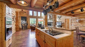 A log cabin interior with a kitchen featuring wooden cabinets, a central island with a stove, hanging pots, and a stone fireplace. - Old Trail Hideaway: 3  BR, 2  BA House in Wilson, Sleeps 9 (Wilson)