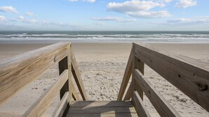 Een houten looppad dat naar een strand leidt, met een heldere lucht en zee op de achtergrond.