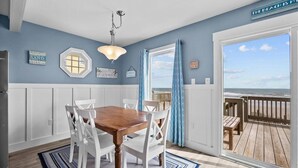 Een eetruimte met een houten tafel en witte stoelen, een hanglamp en uitzicht op het strand door een grote schuifdeur.