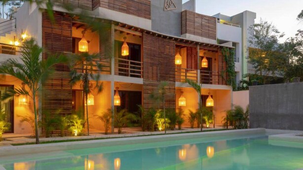 A modern multi-story building with a swimming pool, featuring balconies with wooden railings and warm lighting. - Cozy Rooms to let (Tulum)