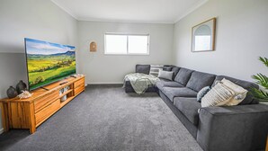 A living room with a grey sofa, a wooden entertainment unit, a flat-screen TV, a window, and a framed artwork on the wall.