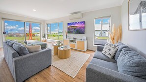 A spacious living room with a grey sofa set, a wooden coffee table, and a large TV mounted on the wall.