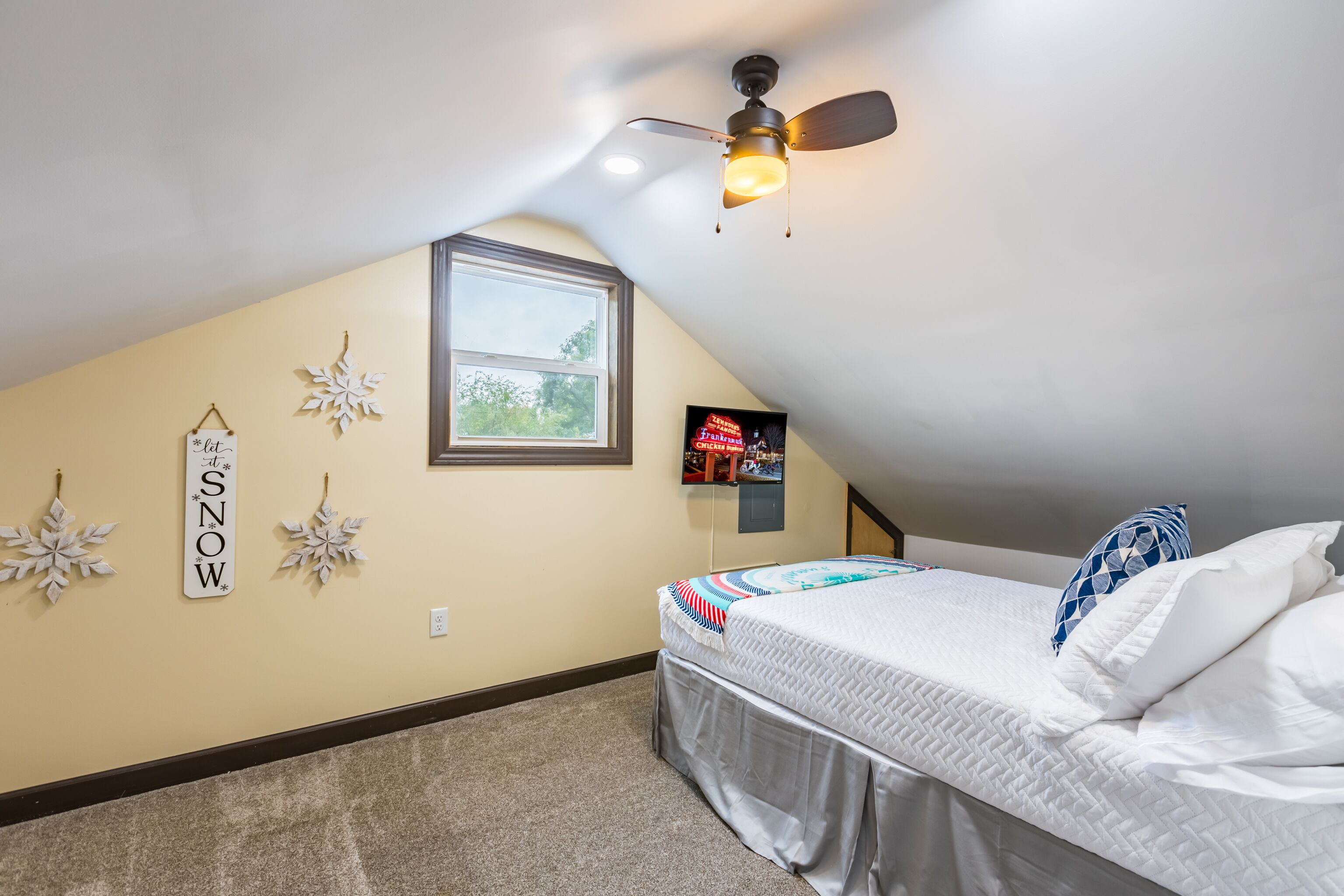 A bedroom with a sloped ceiling, a bed with white bedding, a ceiling fan, a window, and snow-themed wall decorations.