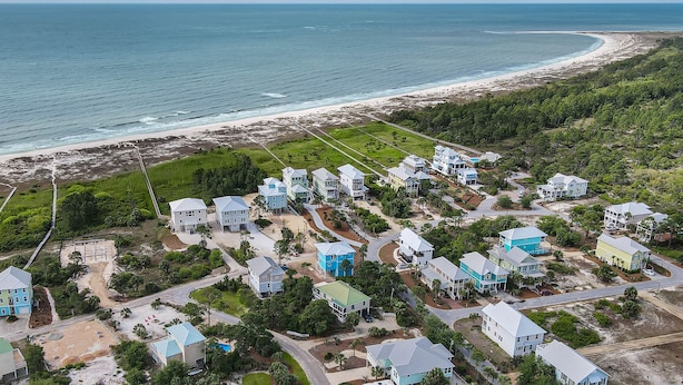 An aerial view of a coastal residential area with houses, a beach, and a road leading to the shore.