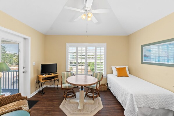 A room with a bed, a TV on a stand, a dining table with chairs, and a ceiling fan.