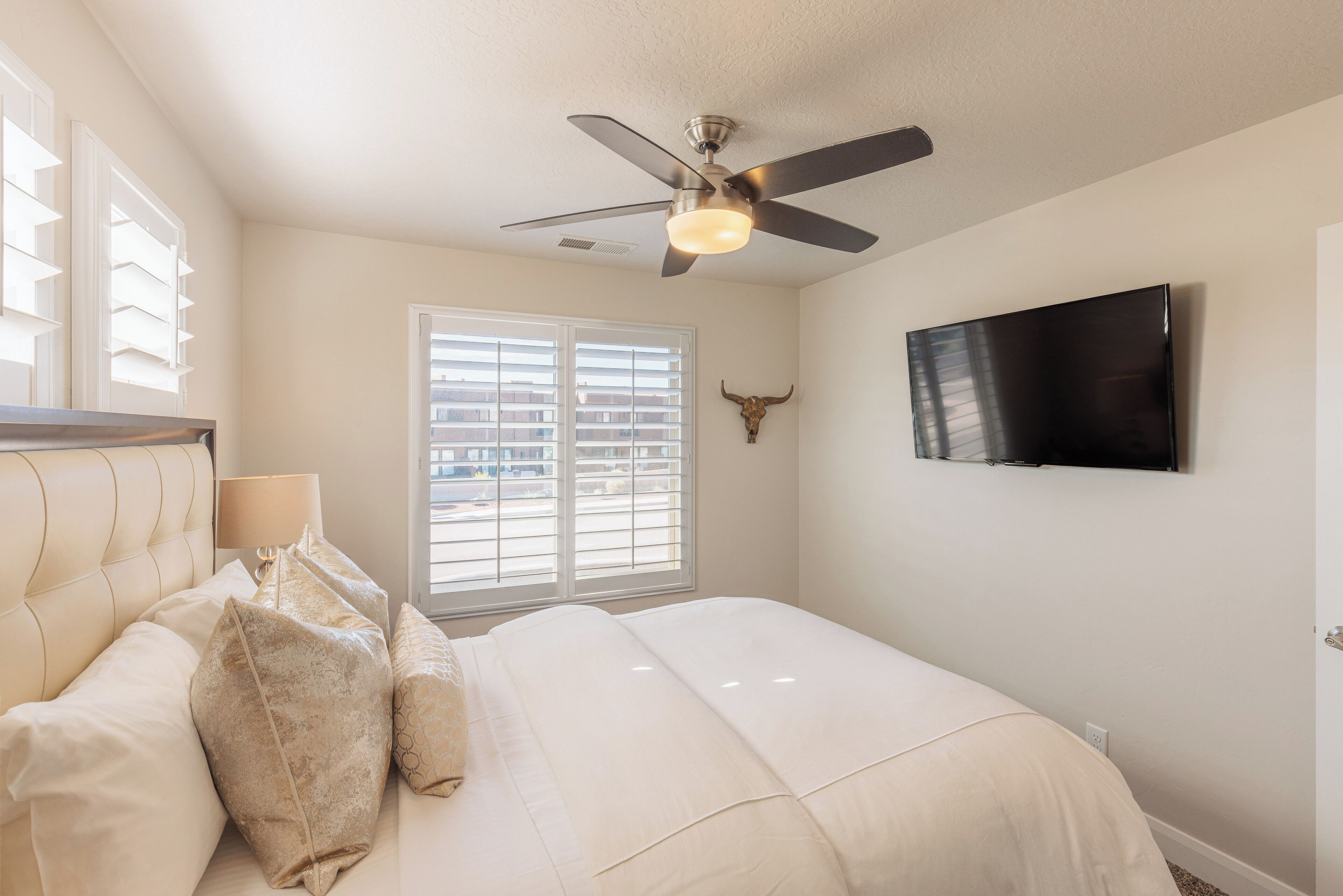 Un dormitorio con cama, ventilador de techo, televisor montado y ventana con persianas.