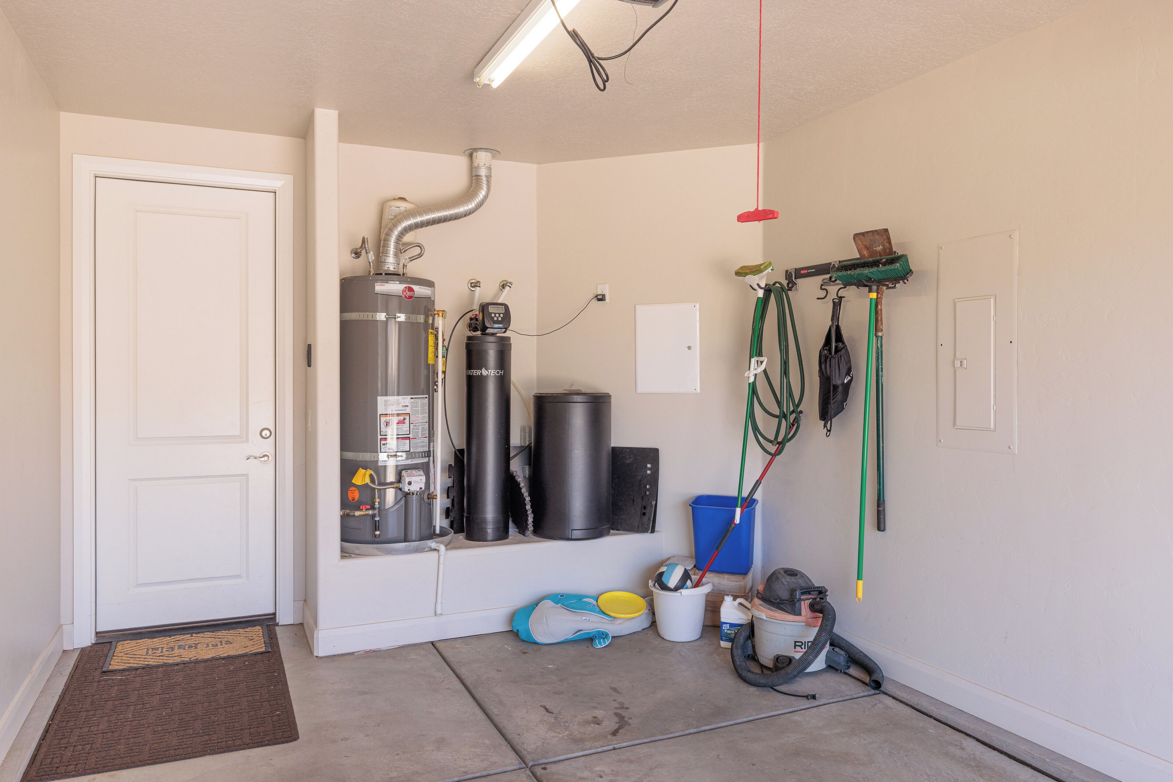A garage with a white door, a water heater, a vacuum cleaner, and a hose.