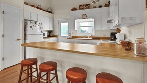 A kitchen with white cabinets, a wooden island with stools, a clock, and a window. - Dream On #145 - Private boat dock, short walk to the beach, dog friendly! (Topsail Beach)