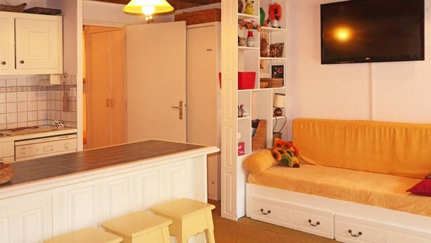 A cozy living room with a sofa, a TV mounted on the wall, and a kitchen area with a dining table and chairs.