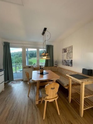 Une pièce comprenant une table à manger en bois, des chaises, un banc, un luminaire suspendu, un radiateur et une vue sur la verdure à l’extérieur.