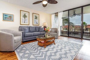 A living room with a glass coffee table, two sofas, a patterned rug, and a view through a glass door to an outdoor area with chairs. - KONA PACIFIC C-618 NICE OCEAN VIEW, Top Floor, WALK TO TOWN! Elevator, AC!! (Kailua Kona)