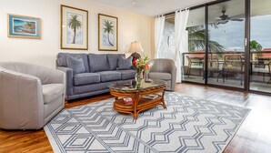 Ein Wohnzimmer mit einem Glastisch, zwei Sofas, einem gemusterten Teppich und Blick durch eine Glastür auf einen Außenbereich mit Stühlen.