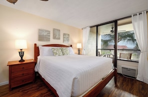 A bedroom with a wooden bed, two bedside tables with lamps, a window with a view of palm trees, and artwork on the wall. - KONA PACIFIC C-618 NICE OCEAN VIEW, Top Floor, WALK TO TOWN! Elevator, AC!! (Kailua Kona)
