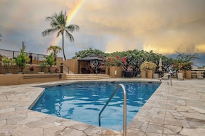 A swimming pool with a metal ladder, surrounded by stone tiles, palm trees, and a gazebo. - KONA PACIFIC C-618 NICE OCEAN VIEW, Top Floor, WALK TO TOWN! Elevator, AC!! (Kailua Kona)