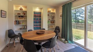 A dining area with a round wooden table surrounded by black chairs, shelves with decorative items, and a view of the outdoors through a sliding glass door.
