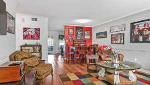 A living room with a sofa, armchair, and a coffee table. There is a dining area with a table and chairs. The walls are decorated with sports memorabilia. - Five Points Dawg Crib On Campus (Athens)