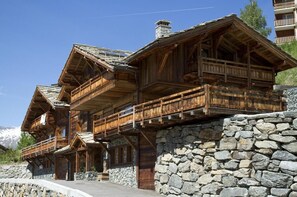 A wooden chalet with balconies, stone walls, and a mountainous backdrop.