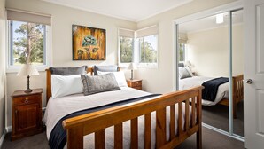 A bedroom with a wooden bed, two bedside tables, a nightstand, a large mirror, and a painting on the wall. - High Peaks Cottages (Fumina South)