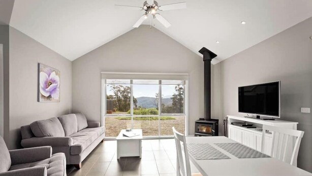 A modern living room with a grey sofa, a white dining table, and a TV. - High Peaks Cottages (Fumina South)