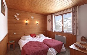 A cozy room with wooden walls, a bed with red and white bedding, a small table with a chessboard, and a window with a view of a snowy landscape.