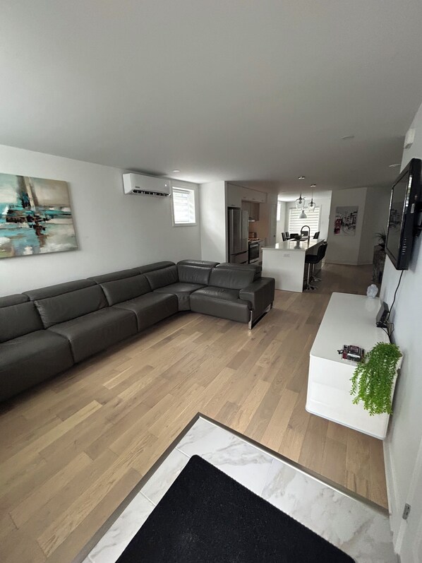 A modern living room with a sectional sofa, a dining area with a white table and chairs, and a kitchen in the background.