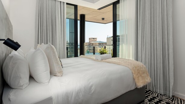 A modern bedroom with a large bed, curtains, and a view of buildings through large windows. - 529 Station House (Cape Town)