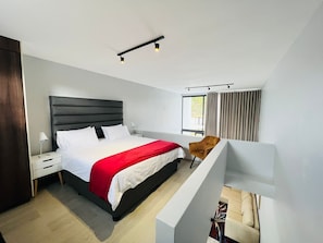 Ein modernes Schlafzimmer mit einem großen Bett, einem Nachttisch, einem Sessel und einem Fenster mit Vorhängen.